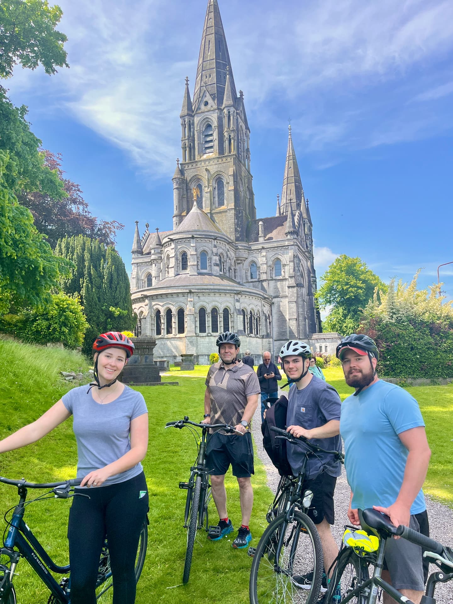Four people standing with their bikes on the grass in front of St Fin Barre's Cathedral in Cork