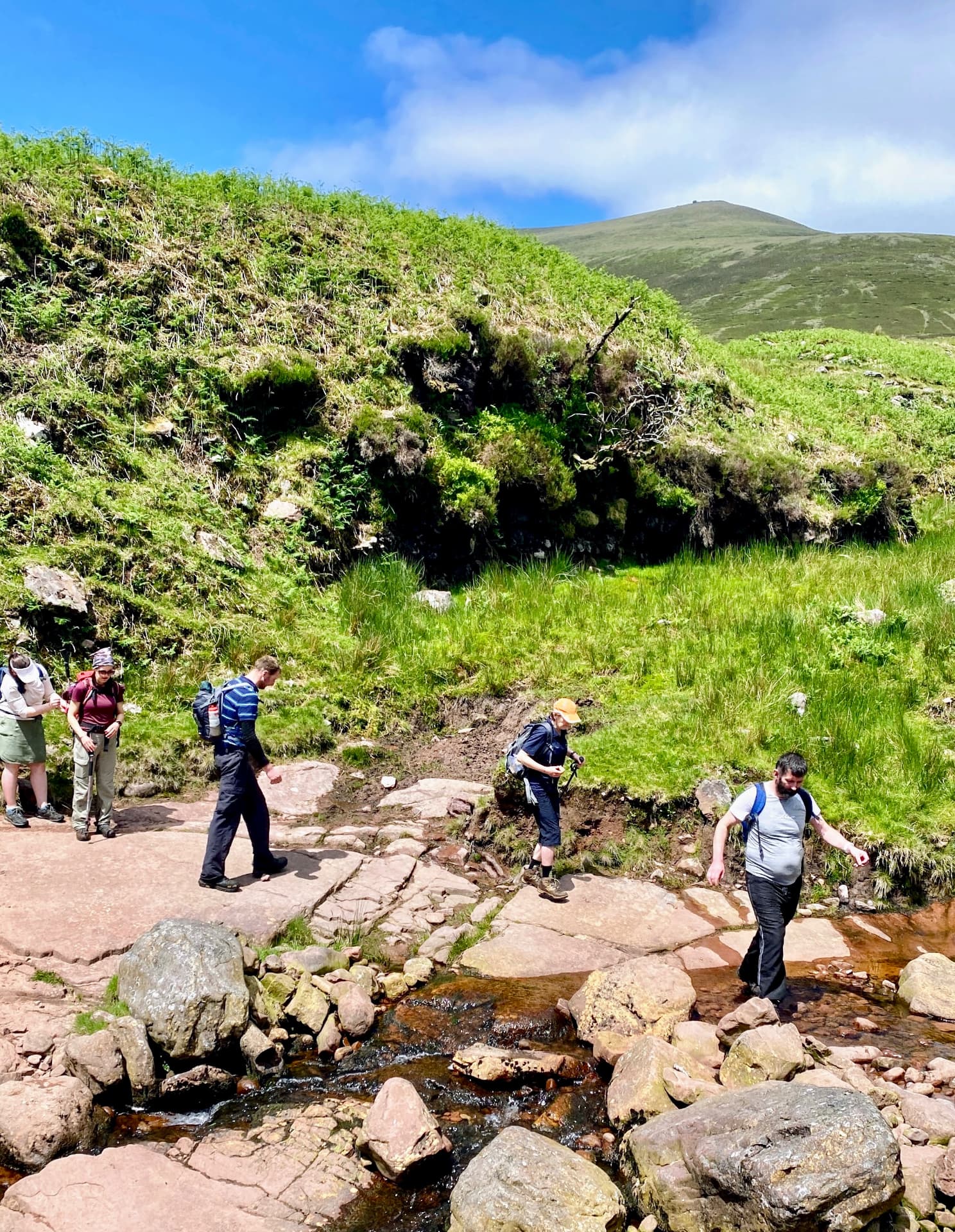 A group of hikers crossing a rocky mountain stream on a sunny day in Munster