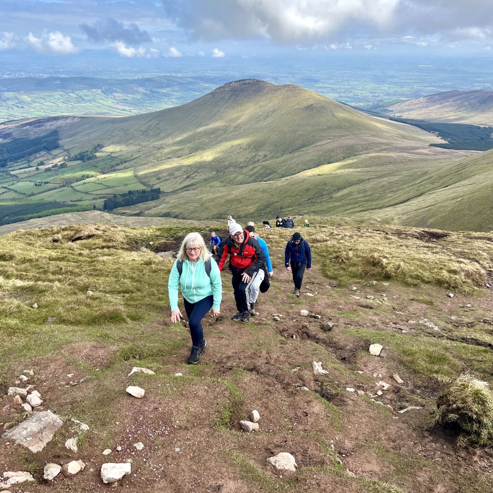 Walkers climbing a grassy mountain path at a relaxed pace with wide countryside views behind them