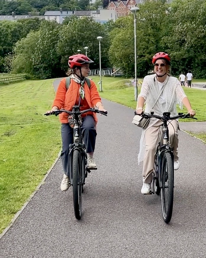 Two women cycling side by side along a path through a green park in Cork