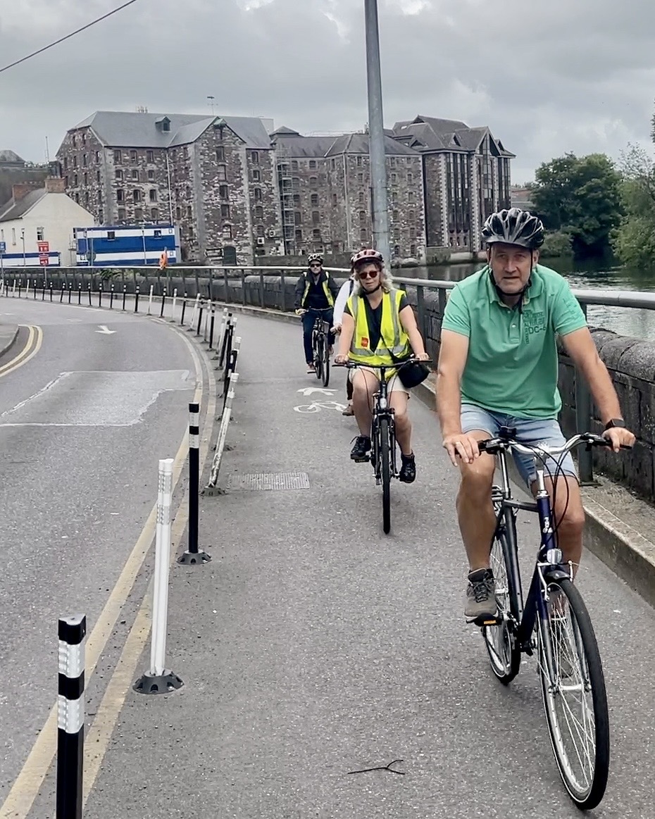 Cyclists riding in single file along a protected cycle lane beside the River Lee, with old Cork warehouse buildings behind