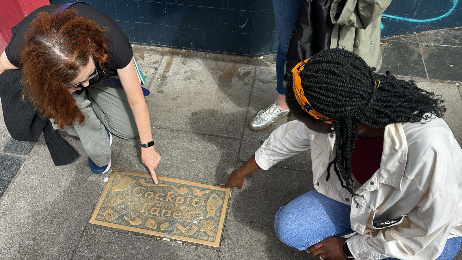 Two participants kneeling to examine a Cockpit Lane pavement plaque while a third looks on, solving a scavenger hunt clue in Cork City