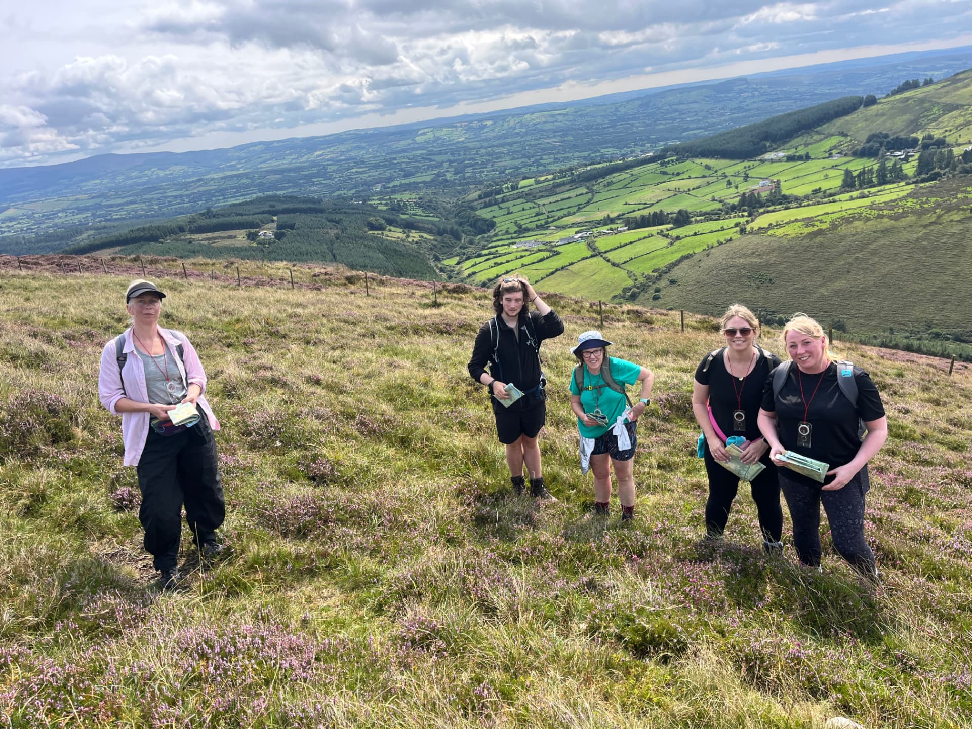 Five Mountain Skills participants standing on a heather hillside holding maps, with green farmland in the valley below