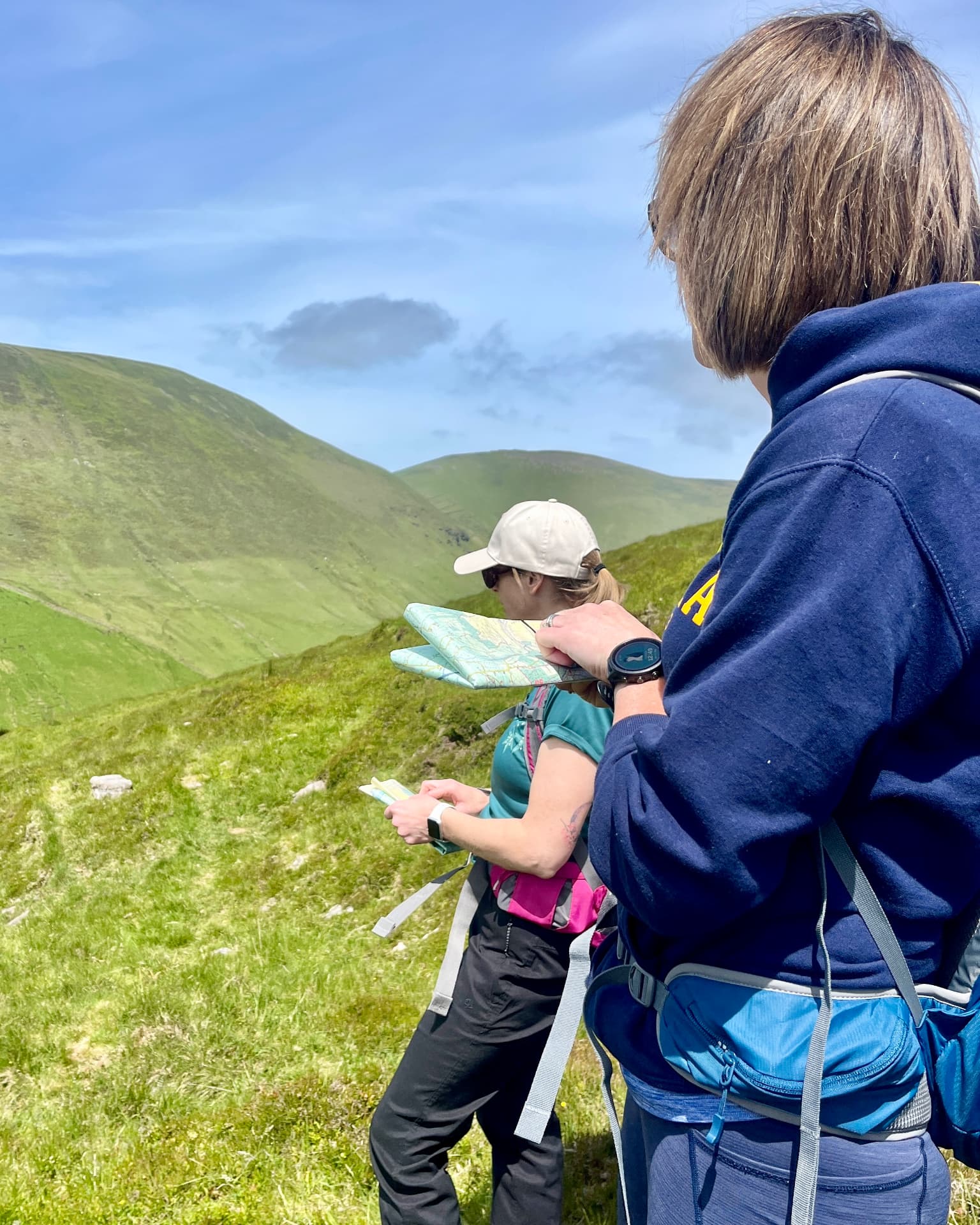 Two Mountain Skills participants holding open maps on a grassy mountain slope, with green ridges and a valley behind them