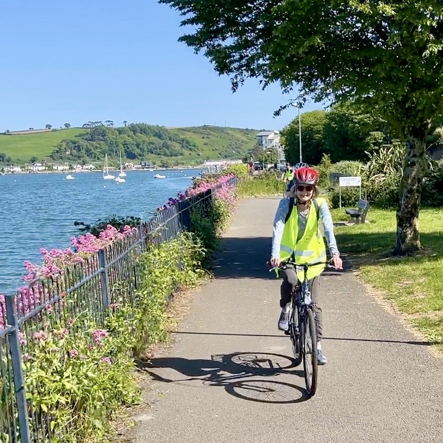 Cyclist on a waterside Greenway path beside flowers, with boats, the harbour and a green hillside behind