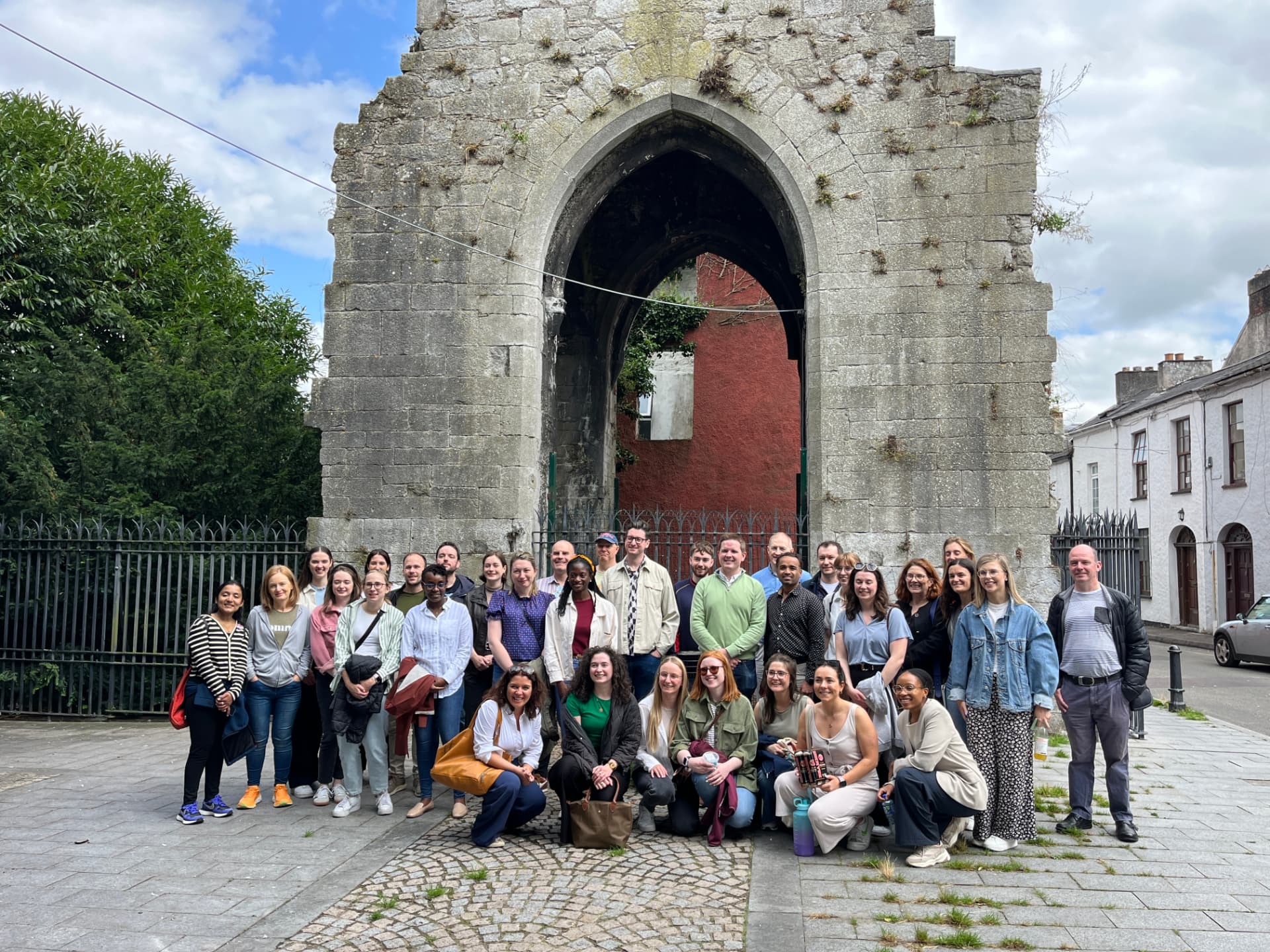 A workplace group of around forty colleagues standing in rows in front of a stone archway in Cork City
